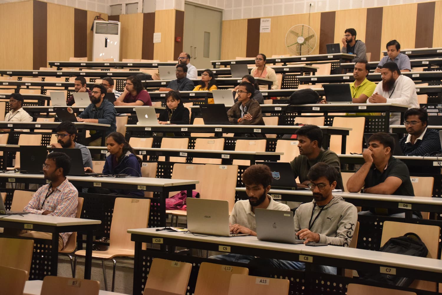 Large auditorium filled with diverse attendees engaged in a workshop session. Participants are seated at tiered desks with laptops open, actively typing and learning during the conference.