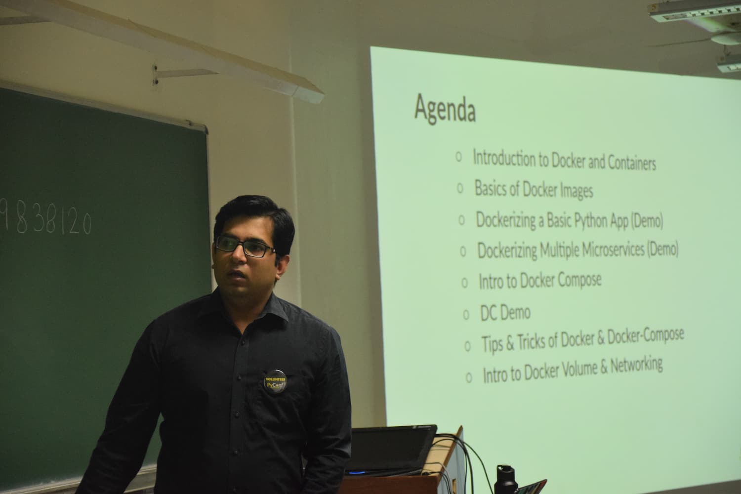 Speaker wearing a PyConf Volunteer badge presenting on Docker. A projection screen shows an agenda for Docker and Docker Compose topics, with a green chalkboard visible in the background.