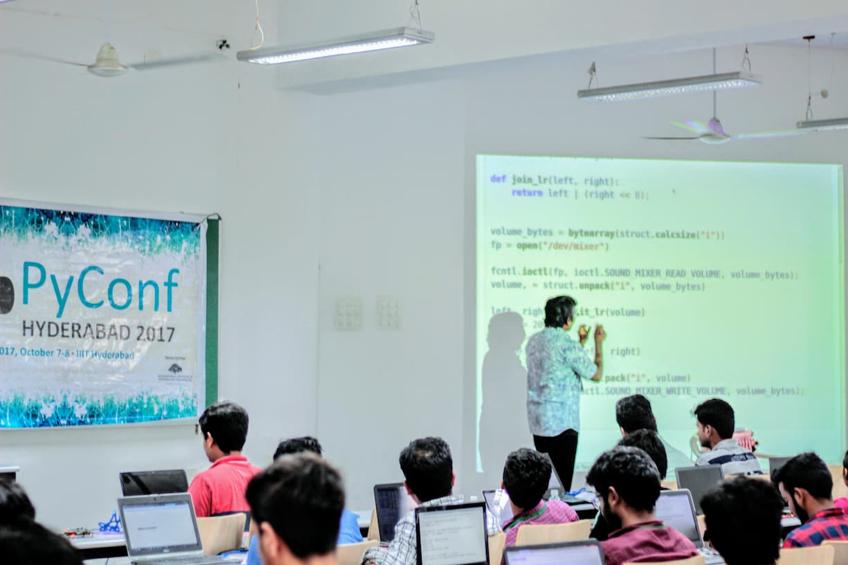 PyConf Hyderabad 2017 coding session with a speaker presenting Python code on a projection screen. Attendees are seated at tables with laptops open, actively coding along during the technical workshop.