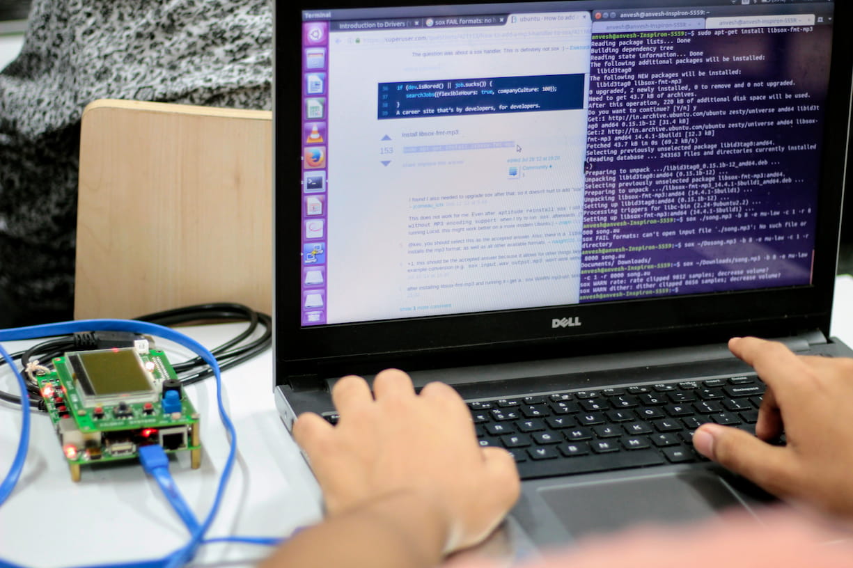 Close-up of a developer working on a Dell laptop displaying a Linux terminal and web browser. A green circuit board connected by blue cables sits on the desk, showing hands-on hardware and software development.