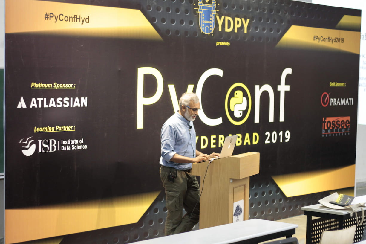 Speaker with gray hair and beard presenting at PyConf Hyderabad 2019, standing at a wooden podium with a laptop. Large black and gold conference banner displays sponsors including Atlassian, ISB, Pramati, and FOSSEE.