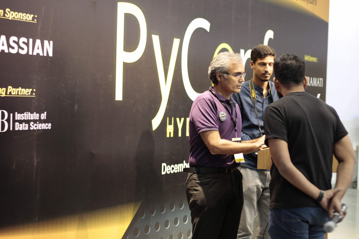 Three men engaged in conversation at PyConf Hyderabad conference. Conference branding banner visible in background showing PyConf HYD with sponsor logos including Atlassian and ISB Institute of Data Science.