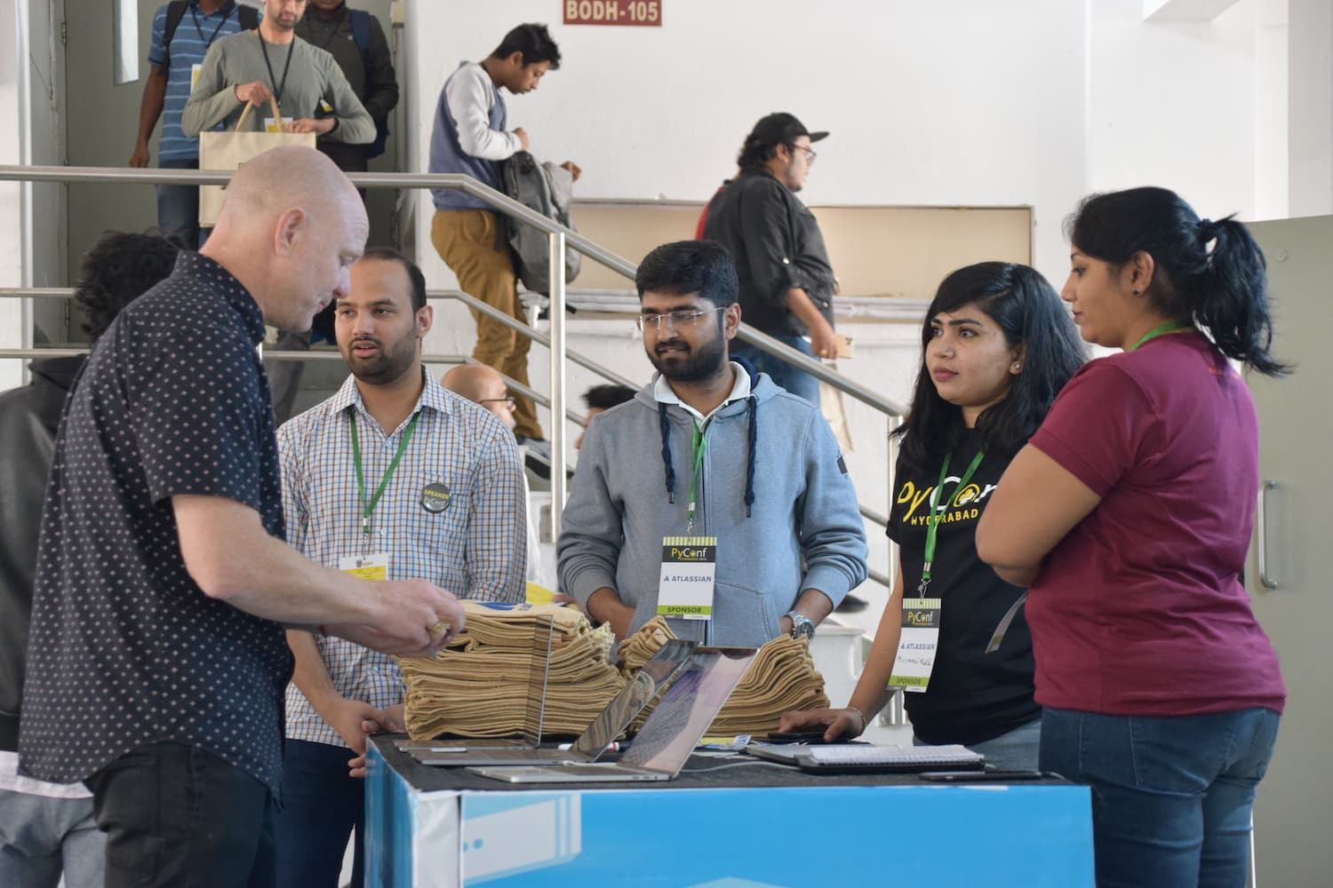 Conference registration desk with organizers assisting attendees. Staff members wearing PyConf Hyderabad t-shirts and Atlassian sponsor badges help participants at the registration counter.