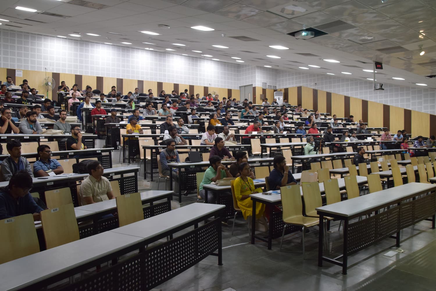 Wide shot of a large auditorium filled with 100-150 attendees during a conference session. Participants are seated at tiered desks, many with laptops open, focused on the presentation.