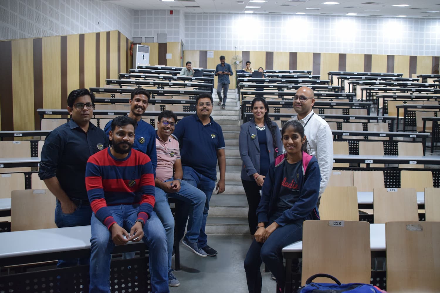 Group of eight smiling organizers and volunteers posing in an empty lecture hall, wearing event badges. They are positioned among rows of light wooden desks and chairs.
