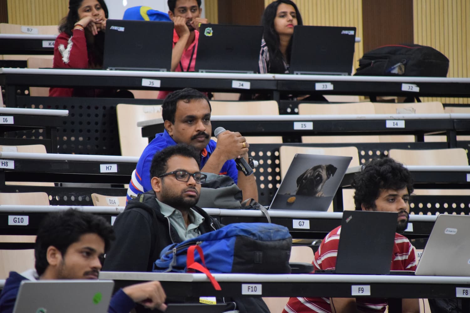 Conference session in progress with a speaker using a microphone. Attendees are seated at desks with laptops open, including one displaying a pug dog image, actively participating in the workshop.
