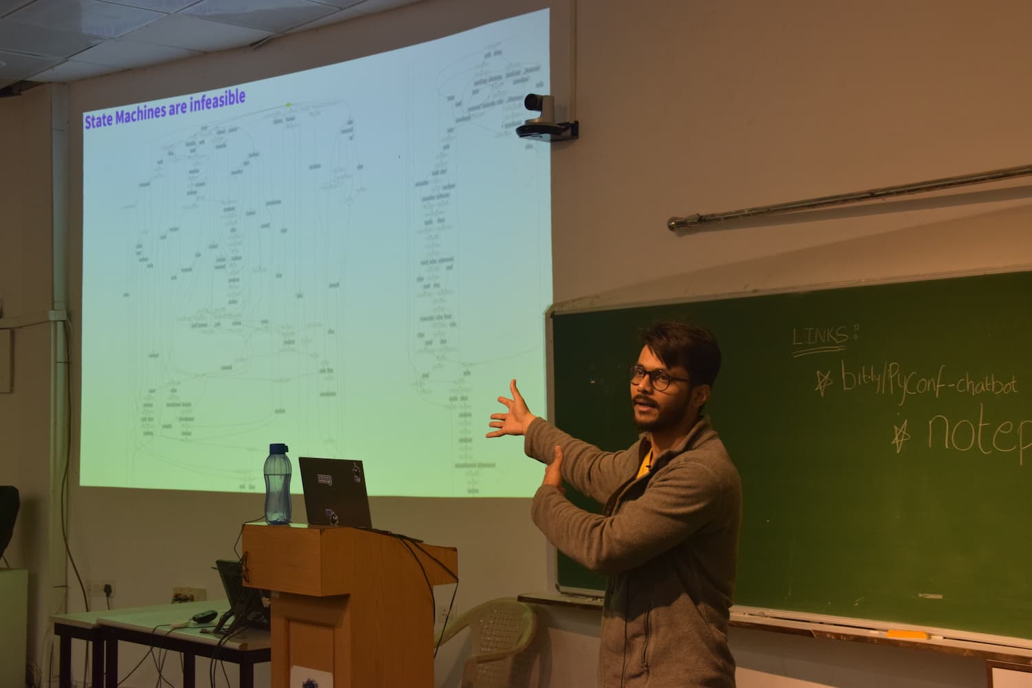 Speaker presenting on State Machines, gesturing in front of a projection screen showing a complex flowchart diagram. A green chalkboard with links is visible on the right side.