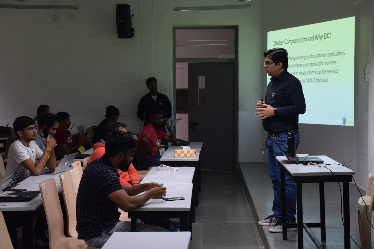 Technical presentation on Docker Compose in a classroom setting. Speaker gestures to an audience of attendees working on laptops at desks, with the presentation slide visible on the projector screen.